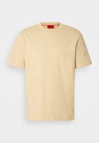 T-shirt beige à manches courtes et col rond avec un logo "HUGO" discret sur le devant et une étiquette intérieure rouge de la marque au niveau du col.