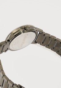 Ρολόι χειρός Calvin Klein από ανοξείδωτο ατσάλι με μπρασελέ από μεταλλικούς συνδέσμους, ορατό κουρδιστήρι και χαραγμένη πίσω πλάκα που δείχνει το εμπορικό σήμα και τις προδιαγραφές.