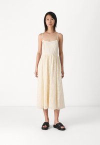 Vestido maxi color crema con tirantes finos, tela texturizada, cuerpo ajustado y falda fluida. Llevado con sandalias de plataforma negras.