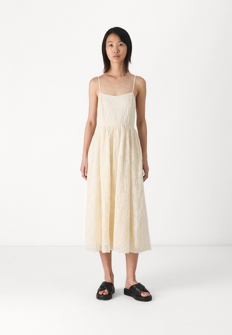 Vestido maxi color crema con tirantes finos, tela texturizada, cuerpo ajustado y falda fluida. Llevado con sandalias de plataforma negras.