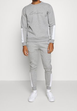 Mand iført en grå sweatshirt og matchende joggingbukser med hvide striber på ærmer og ben, kombineret med hvide sneakers.
