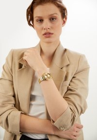 Reloj de pulsera dorado con una pulsera gruesa de eslabones, esfera redonda y acabado pulido. Lleva puesto en la muñeca de un modelo, combinado con una chaqueta beige.