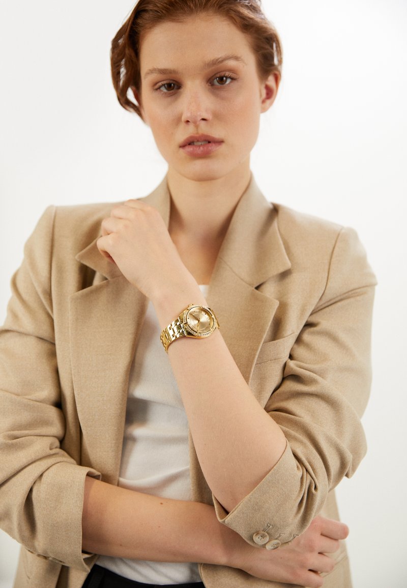 Reloj de pulsera dorado con una pulsera gruesa de eslabones, esfera redonda y acabado pulido. Lleva puesto en la muñeca de un modelo, combinado con una chaqueta beige.