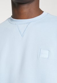 Ljust blå sweatshirt i mjukt tyg, med rund hals, strukturerad sömnad och en liten präglad vit fyrkantig logotyp på bröstet.