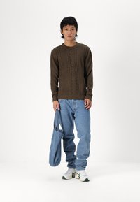 Suéter de punto marrón, jeans azul relajados y zapatillas blancas con acentos verdes; sosteniendo un bolso tote azul. Capa simple y texturizada.