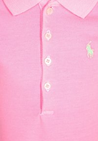 Vaaleanpunainen polo-paita, jossa on neljä nappia ja pieni vihreä brodeerattu hevosen logo vasemmalla puolella rintaa.