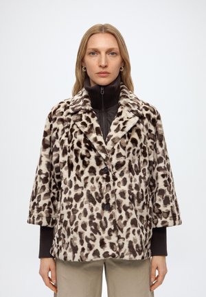 Femme portant un manteau en fausse fourrure à imprimé léopard sur un pull zippé noir et un pantalon beige, debout devant un fond clair uni.