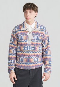 Νέος άνδρας που φοράει φλις πουλόβερ με σχέδια, διαθέτει πατιλέτα με κουμπιά τύπου σναπ και τσέπη στο στήθος με φερμουάρ, συνδυασμένο με σκούρο παντελόνι, στέκεται μπροστά σε λευκό φόντο.