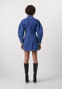 Denim suknelė su prigludusia siluetu, pūstomis ilgomis rankovėmis ir dviem priekinėmis kišenėmis. Medžiaga yra tamsiai mėlynos spalvos su matomais siuvimo detalėmis.