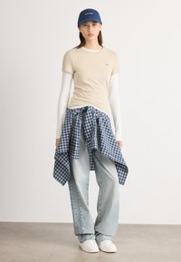 Beige t-skjorte med korte ermer lagd over en hvit skjorte med lange ermer, blå rutete skjorte knyttet rundt livet, lyseblå vid bukse, hvite joggesko, blå caps.