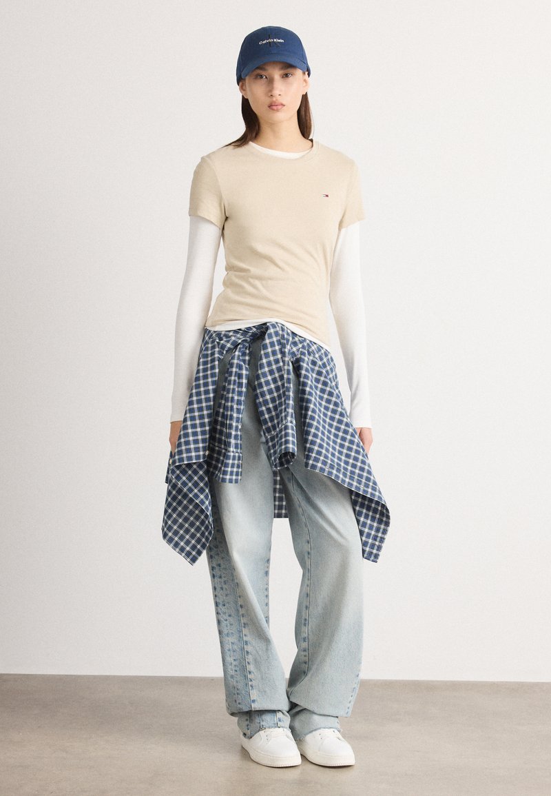 Beige t-skjorte med korte ermer lagd over en hvit skjorte med lange ermer, blå rutete skjorte knyttet rundt livet, lyseblå vid bukse, hvite joggesko, blå caps.