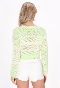 Grüner, gestrickter, verkürzter Pullover mit Lochmuster und gewellten Säumen. Kombiniert mit weißen, ausgefransten Jeansshorts.