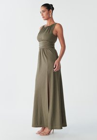 Das Modell in einem olivgrünen, bodenlangen Kleid mit einem Seitenschlitz posiert elegant, trägt Ohrringe mit Ohrhängern und beigefarbene Sandalen mit Riemchen vor einem schlichten Hintergrund.