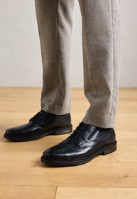 Zapatos de vestir de cuero negro con cordones, llevados con pantalones de traje gris claro sobre un suelo de madera.