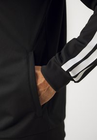 Main dans la poche d'une veste noire avec des rayures blanches sur la manche, sur un fond clair uni.