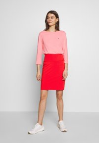 Maglia rosa a maniche lunghe e gonna a tubino rossa, abbinate a sneaker bianche. L'outfit presenta una silhouette aderente e una texture liscia.