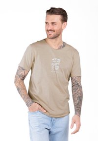Hombre con tatuajes en ambos brazos, vistiendo una camiseta gráfica beige y jeans de color azul claro, sonriendo y mirando de lado con una mano en el bolsillo.