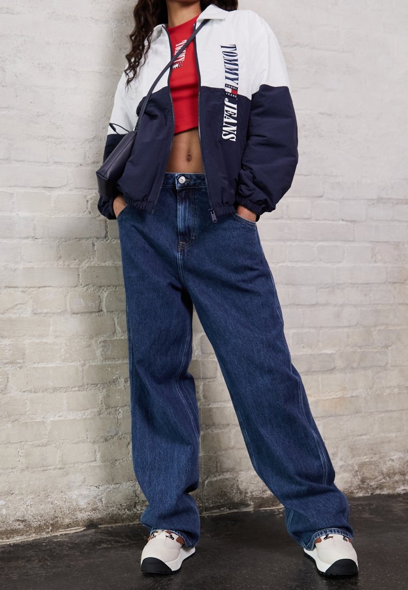 Navy and white color-blocked bomber jacket with logo, red crop top, loose blue jeans, and beige sneakers with patterned accents.