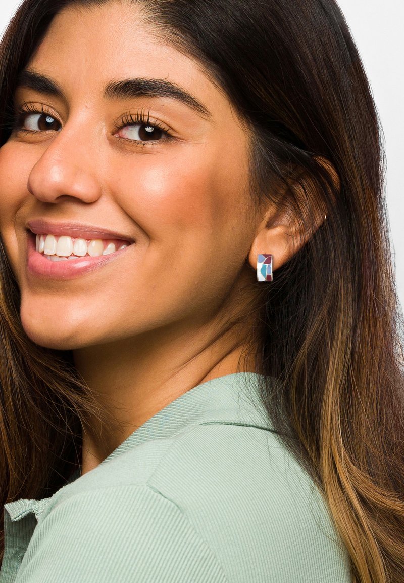 Srebrne geometryczne kolczyki z niebieskimi, białymi i różowymi akcentami, noszone przez osobę z długimi, ciemnymi włosami i miętową koszulą.