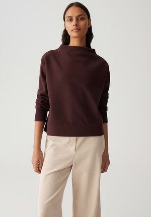 Femme portant un pull à manches longues marron foncé avec un col haut et un pantalon beige à larges jambes en côtes, se tenant devant un fond uni.