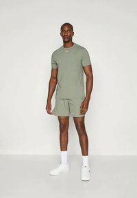 Olivengrønn treningssett som inkluderer en tettsittende T-skjorte med korte ermer og matchende korte bukser, laget av glatte stoffer, kombinert med hvite sneakers.