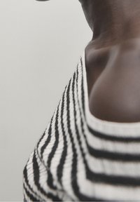 Närbild av en persons nacke och axel som bär en svart- och vitrandig skjorta mot en enkel ljus bakgrund.