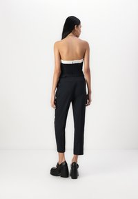 Svart strapless topp med teksturert liv, parret med svarte tapered bukser og klumpete hæler. Modell som står med ryggen til, minimalistisk bakgrunn.