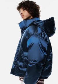 Kurtka puchowa wykonana z błyszczącego niebieskiego materiału, z wysokim kołnierzem, odpinanym kapturem oraz luźnym krojem, z pikowanymi sekcjami i elastycznymi mankietami.