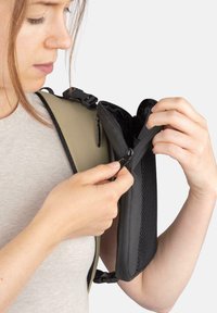 Schwarze Stofftasche, die aufgezogen wird, befestigt an einem beigen Rucksack. Hat eine strukturierte Außenfläche und einen Clipverschluss. Sichtbare Haut mittleren Hauttons.