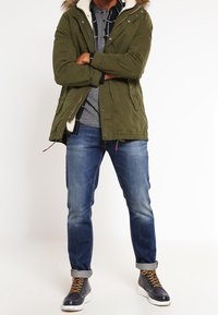 Olivgrön parkas med syntetisk päls huva, texturerat tyg. Grå skjorta i lager under, i kombination med blå jeans och svarta stövlar med gula snören.