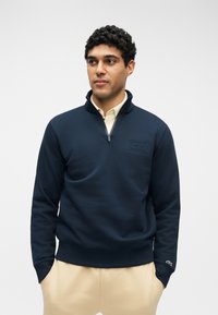 Άνδρας που φοράει μπλε σκούρα μπλούζα με φερμουάρ έως τον λαιμό, με τα χέρια στις τσέπες μπεζ παντελονιού, κοιτάζοντας ελαφρώς προς το πλάι σε απλό φόντο.