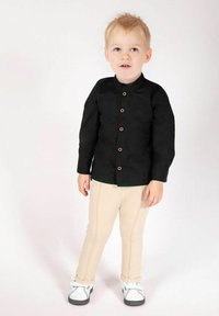 Czarna koszula zapinana na guziki z kołnierzem typu stójka, beżowe dopasowane spodnie i białe buty. Drewniane guziki i gładka faktura tkaniny.