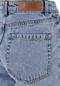 Jean en denim bleu clair avec une étiquette en cuir portant la mention "URBAN CLASSICS DENIM FACTORY." Coutures apparentes et détails de la poche arrière visibles.