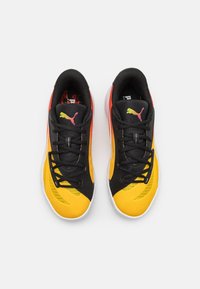 Sorte og gule atletiske sko med en glat tekstur, der har et sort snøresystem, rosa logo-detaljer og en let hvid sål.