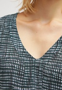 V-paitainen toppi musta-tealraitaisella kuviolla, jossa on vaakasuoria ja pystysuoria viivoja, hienovaraisella tekstuurilla ja poimuilla.