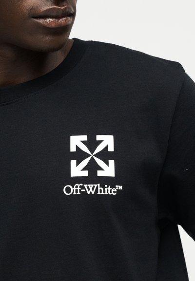 Κοντινή λήψη ενός ατόμου που φοράει μαύρο πουκάμισο με λευκό λογότυπο Off-White που περιλαμβάνει τέσσερα βέλη που δείχνουν προς τα έξω.