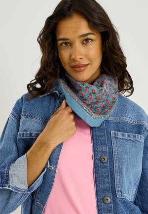 Mujer con el pelo oscuro y ondulado que lleva una chaqueta vaquera, camisa rosa y un pañuelo azul con flores alrededor del cuello, sujetando el pañuelo con una mano.