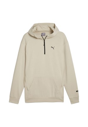 Mężczyzna w jasnobeżowej bluzie z kapturem i pasujących spodniach, stojący tyłem do kamery na jednolitym szarym tle.