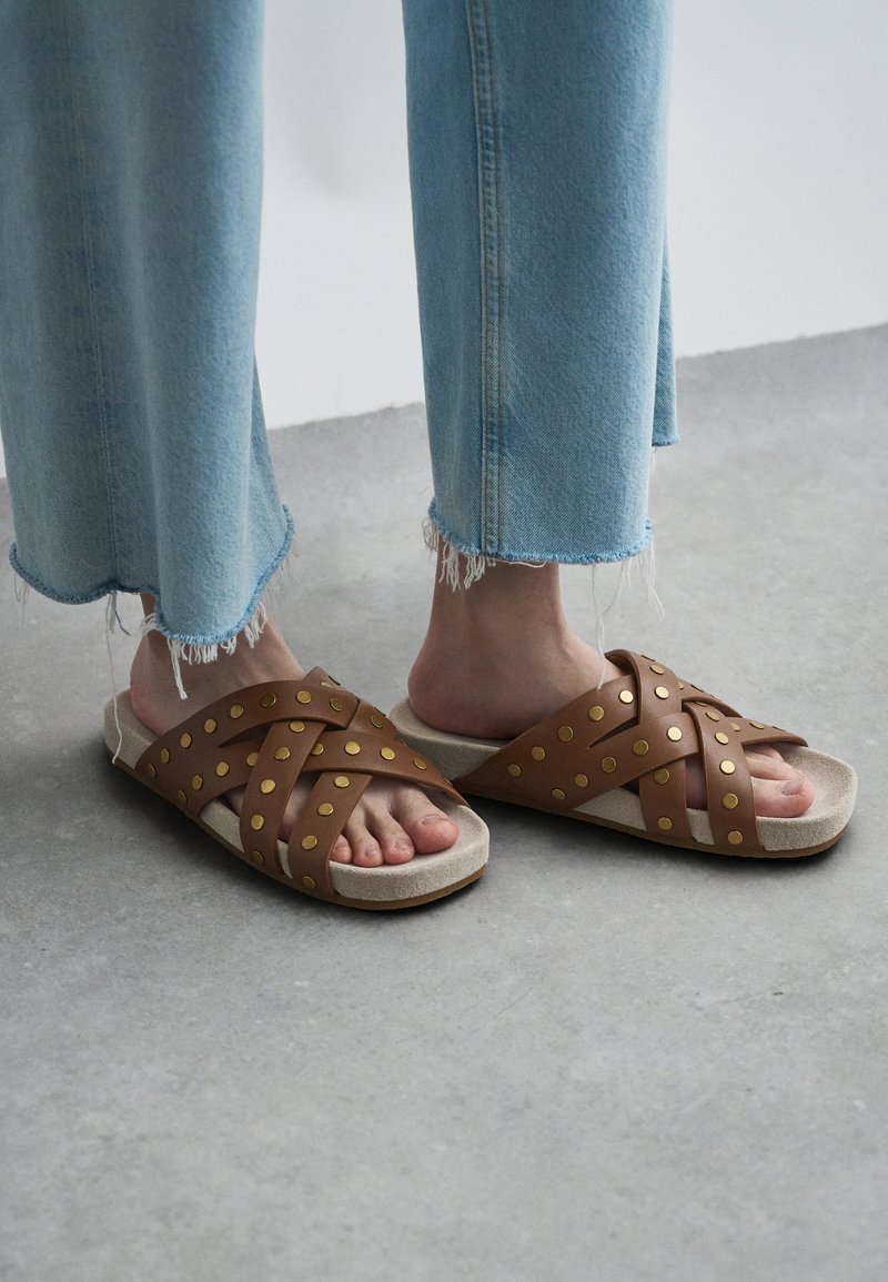 Καφέ σανδάλια με υφασμάτινες σταυρωτές ρίγες, διακοσμημένα με χρυσές τρουκ. Απαλή μπεζ σόλα που συμπληρώνει το σχέδιο. Ορατά τζιν με φθαρμένο στρίφωμα σε μπλε απόχρωση.