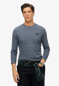 Lange mouwen blauwe heathered shirt met ronde hals, voorzien van een klein logo op de borst, gecombineerd met donkere jeans en een groen geruit flanel.