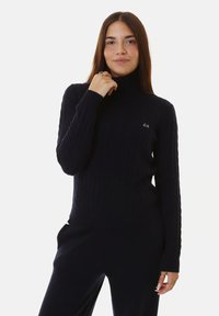 Giovane donna con lunghi capelli castani, che indossa un maglione a collo alto nero a trecce e pantaloni neri coordinati, che posa con una mano vicino al collo.