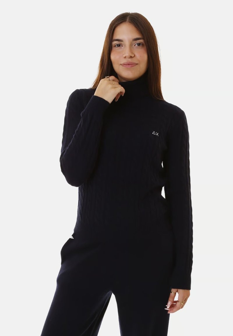 Giovane donna con lunghi capelli castani, che indossa un maglione a collo alto nero a trecce e pantaloni neri coordinati, che posa con una mano vicino al collo.