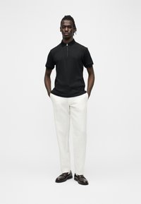 Homme debout, les mains dans les poches, portant un polo noir à manches courtes avec zip, un pantalon blanc et des chaussures formelles marron foncé sur un fond blanc uni.