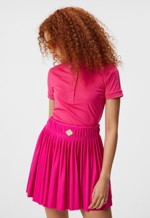 Kobieta z kręconymi czerwonymi włosami, ubrana w różową koszulkę polo i plisowaną różową spódnicę, patrzy w dół i na bok na jednolitym tle.