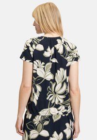 Bloemtop met een marineblauwe achtergrond en grote witte en olijfgroene bloemen. Zachte stof, korte mouwen en een geplooide halslijn.