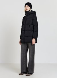 Giacca a vento nera con cappuccio, caratterizzata da sezioni compresse e una zip frontale. Abbinata a pantaloni larghi grigi e scarpe nere.