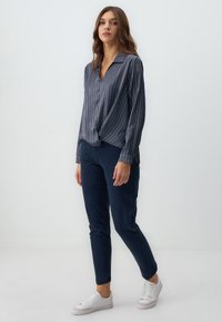 Camicia a righe blu navy e bianca con nodo all'orlo, abbinata a pantaloni blu navy slim-fit e sneakers bianche su uno sfondo neutro.