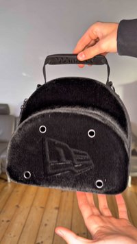 Svart, pälsliknande väska med rundad topp, svart handtag och silverfärgade öljetter. Har präglad design på framsidan och platt botten.