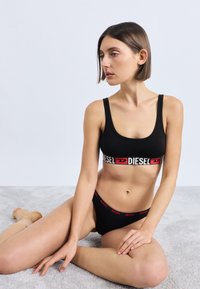 Must spordirinnahoidja ja sobivad tangad punaste "DIESEL" logoga paeladega. Sile kangas, lahtine kaelus ja keha järgiv disain.