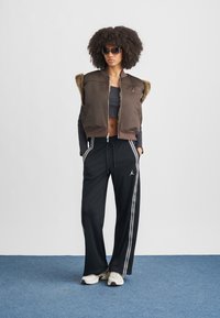 Καφέ γιλέκο με φερμουάρ και επένδυση από ψεύτικη γούνα, μαύρο κροπ τοπ, μαύρο αθλητικό παντελόνι με λευκές ρίγες και λευκά αθλητικά παπούτσια. Γυαλιά ηλίου συμπληρώνουν το look.